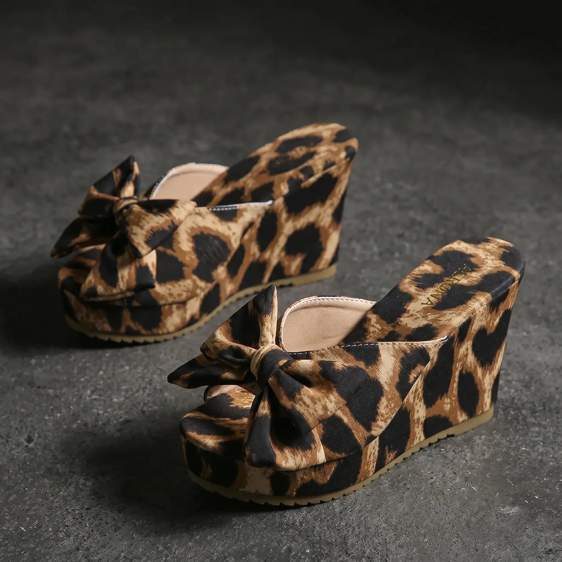 Aerona leopard print wedge sandals featuring an oversized bow detail, peep-toe, and fabric upper, shown from a three-quarter angle on a grey studio background.
