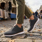 Aerostride breathable knit sneakers in dark grey worn with chinos on a cobblestone street, showing the white EVA sole and brown leather accents.