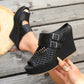 Black Aethel woven wedge sandals with peep toe and dual buckle straps shown on a woman's foot against a wooden background, with a straw hat nearby.