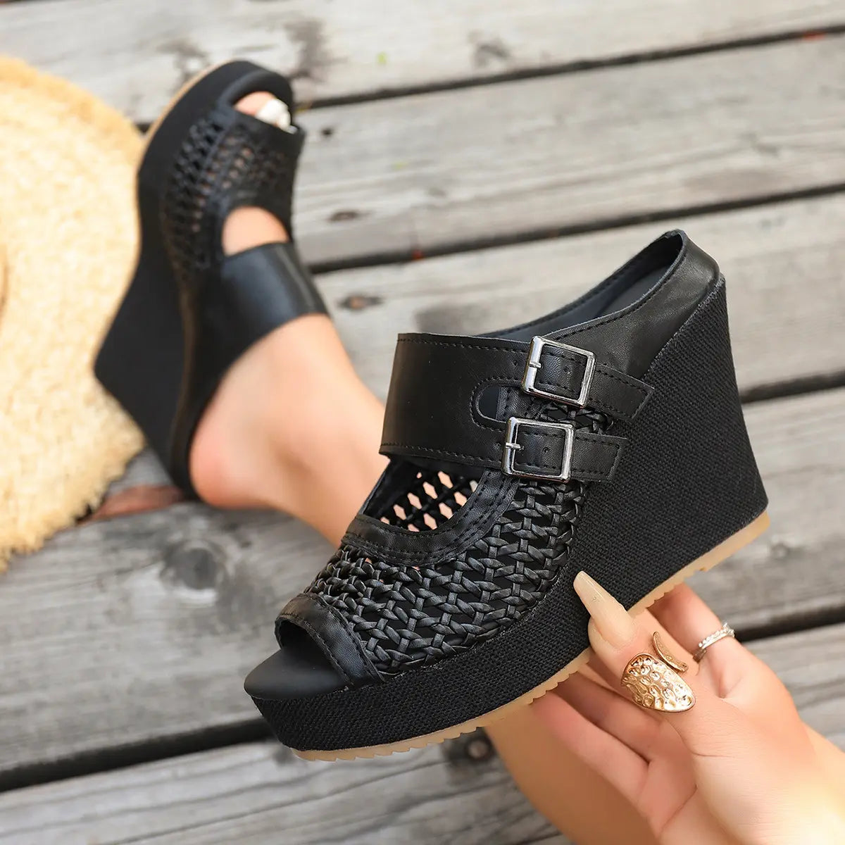 Black Aethel woven wedge sandals with peep toe and dual buckle straps shown on a woman's foot against a wooden background, with a straw hat nearby.