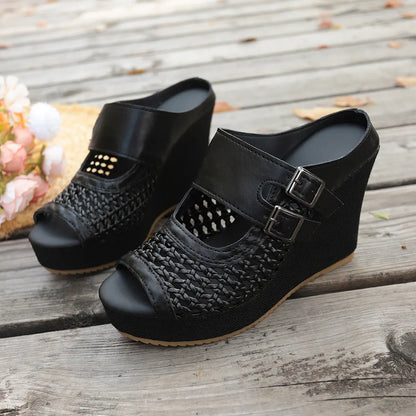 A pair of black Aethel women's woven wedge sandals with peep-toe fronts and dual buckle straps, showcased on a rustic wooden deck with decorative flowers and a straw hat.