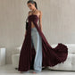 A woman models a deep chocolate brown, flowing maxi dress with a halter neckline and dramatic high front slit, styled stylishly over light-wash wide-leg jeans and sandals, set against a chic interior with a white sofa.