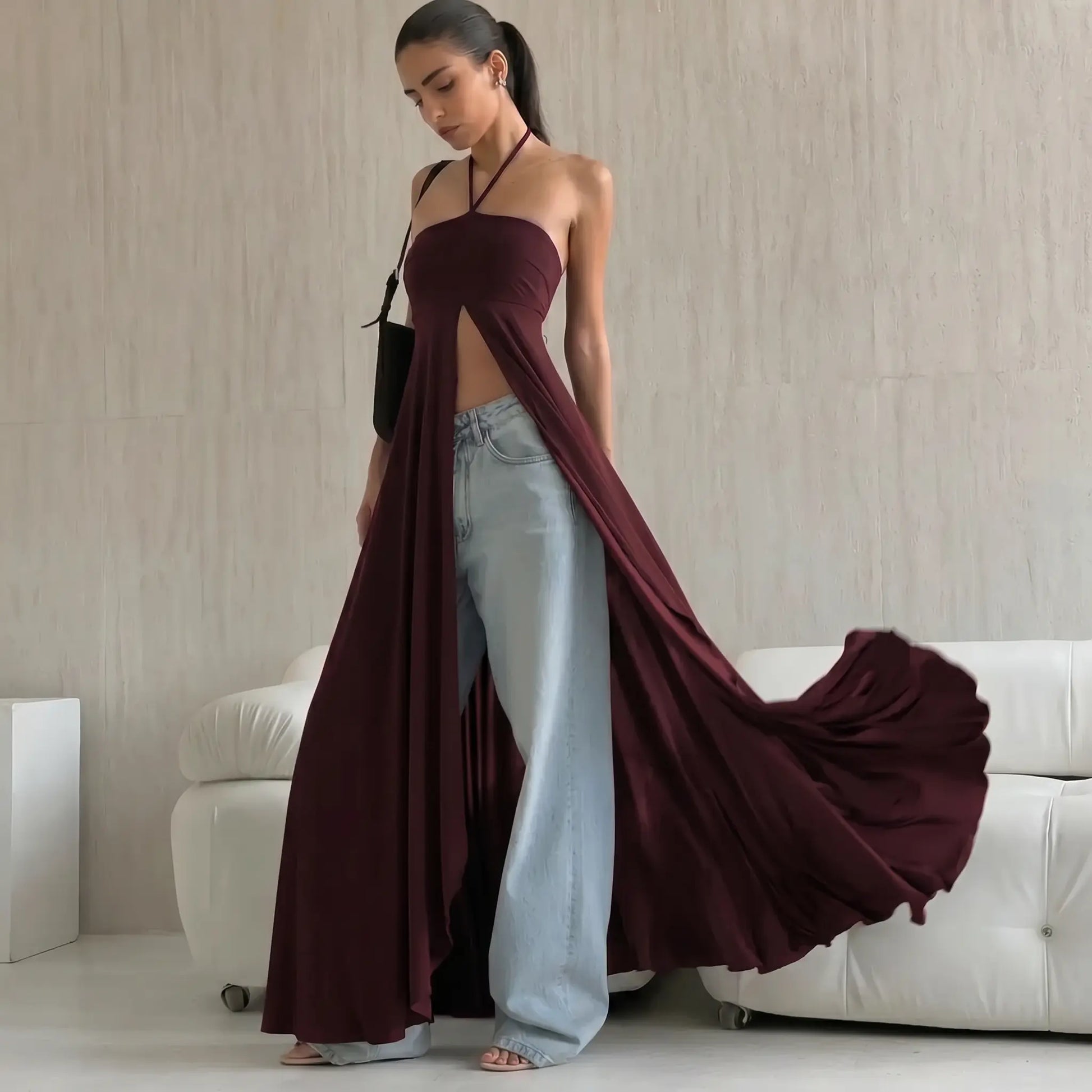 A woman models a deep chocolate brown, flowing maxi dress with a halter neckline and dramatic high front slit, styled stylishly over light-wash wide-leg jeans and sandals, set against a chic interior with a white sofa.