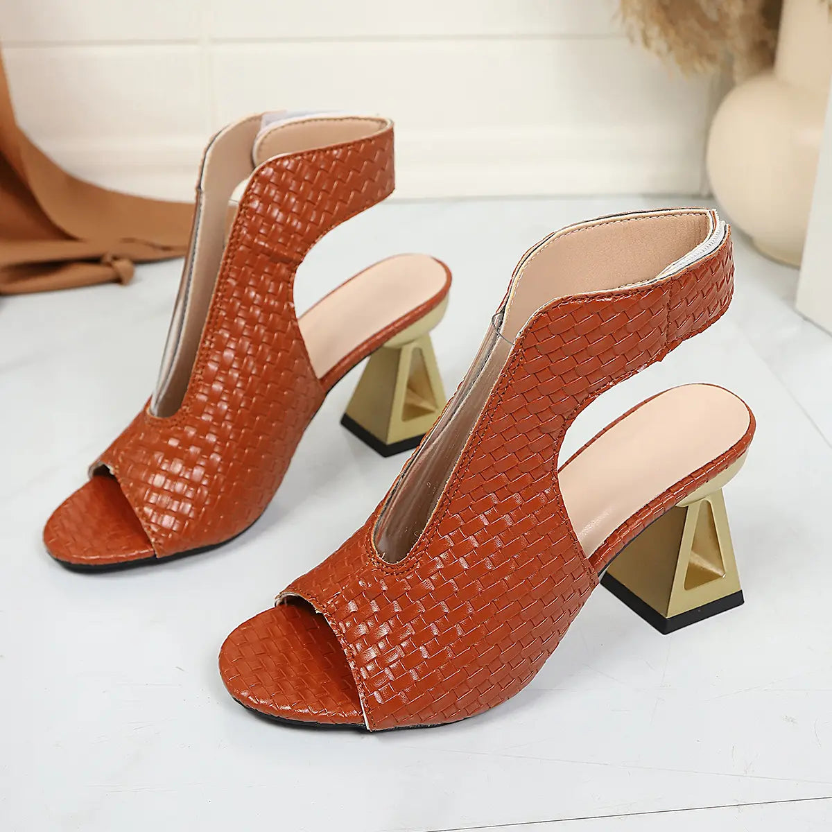 Aethel women's peep-toe block heel sandals in rich cognac brown, featuring a distinctive geometric heel and intricately woven faux leather vamp with a metallic chain ankle strap.