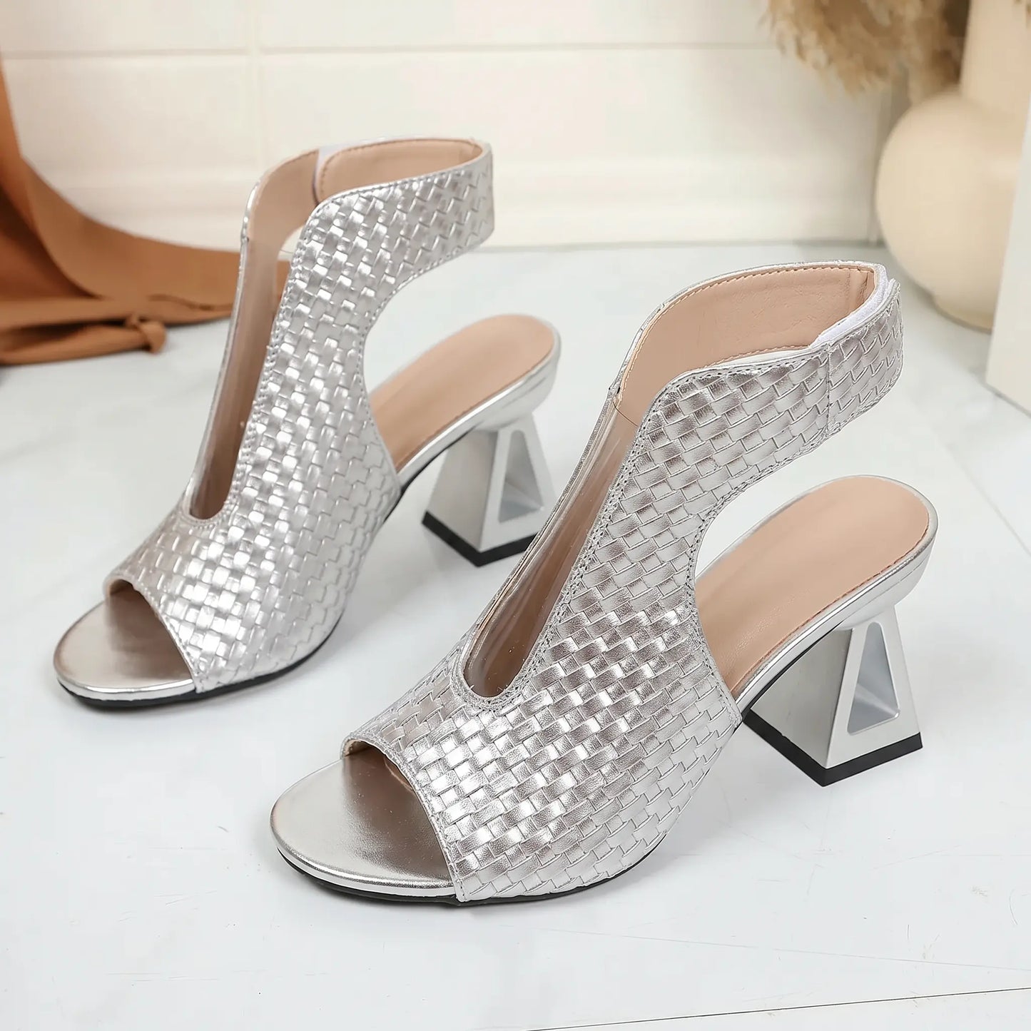 A pair of Aethel brushed silver peep-toe sandals featuring a woven textured upper, a bold geometric block heel, side cut-outs, and a metallic chain ankle strap.