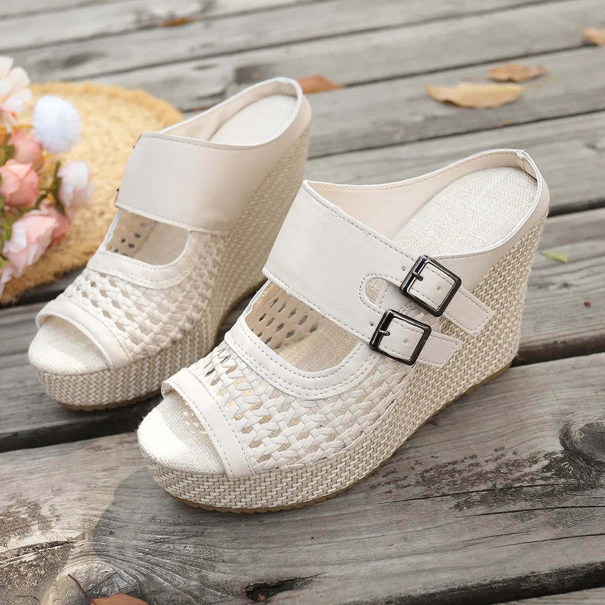 Cream Aethel woven wedge sandals with peep toe and dual buckle straps, displayed on a rustic wooden deck with a straw hat and flowers in the background.