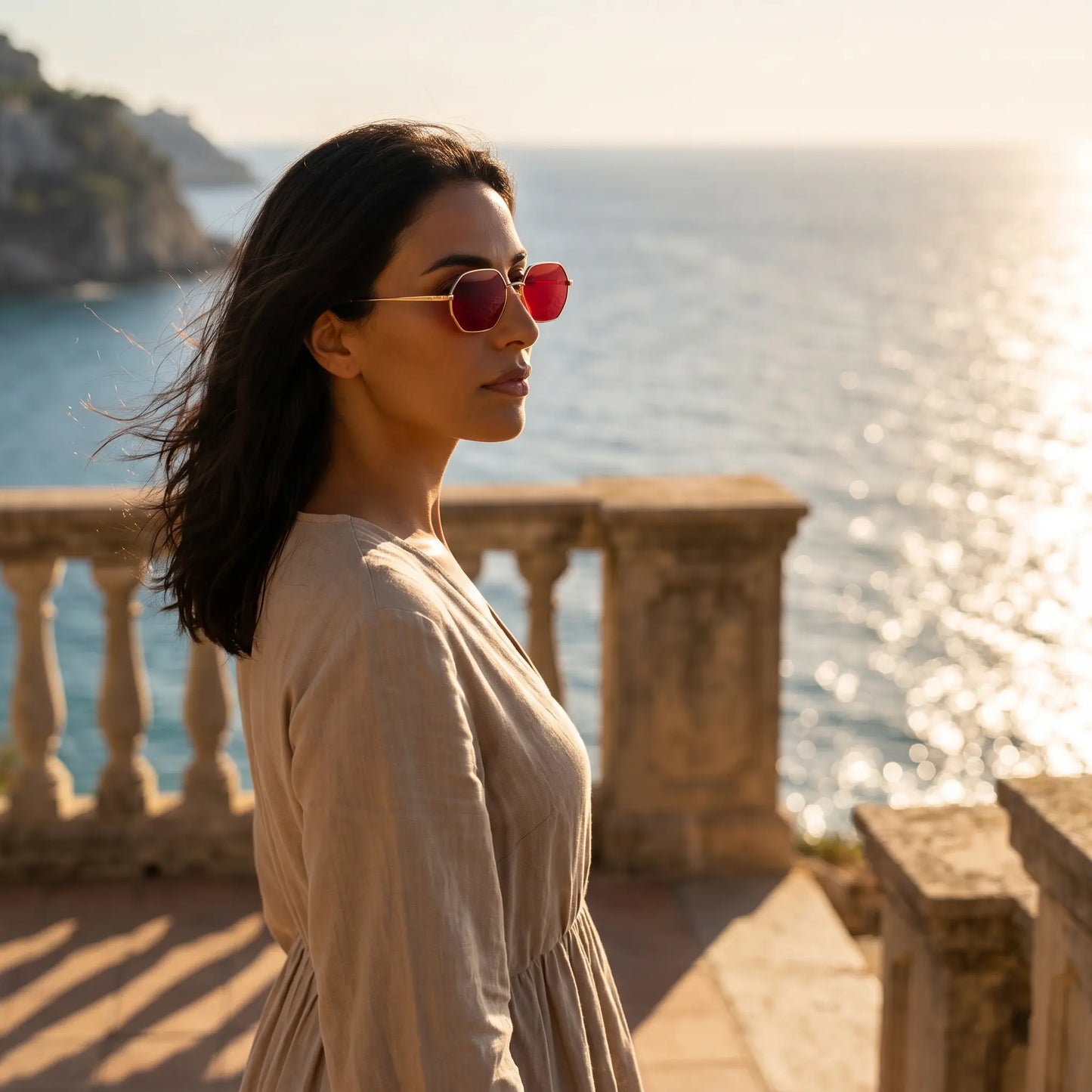 A woman wearing elegant Aethel gold-tone octagonal sunglasses with red lenses, posed by a Mediterranean sea view during golden hour.