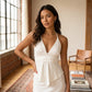 Model wearing an off-white viscose-blend halter neck camisole with a deep V-neck, cinched peplum waist, and asymmetrical hem, styled in an apartment setting with natural light.