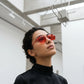 A woman wearing elegant, octagonal sunglasses with vibrant red lenses and gold-tone metal frames, looking up in a minimalist architectural setting.