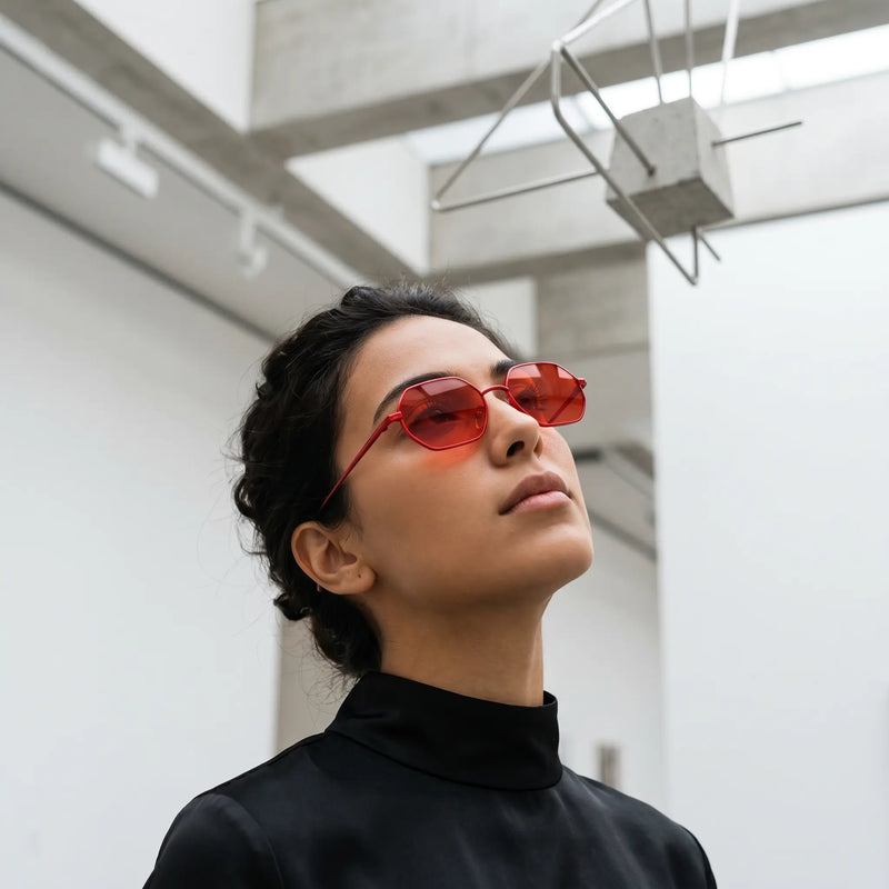 A woman wearing elegant, octagonal sunglasses with vibrant red lenses and gold-tone metal frames, looking up in a minimalist architectural setting.