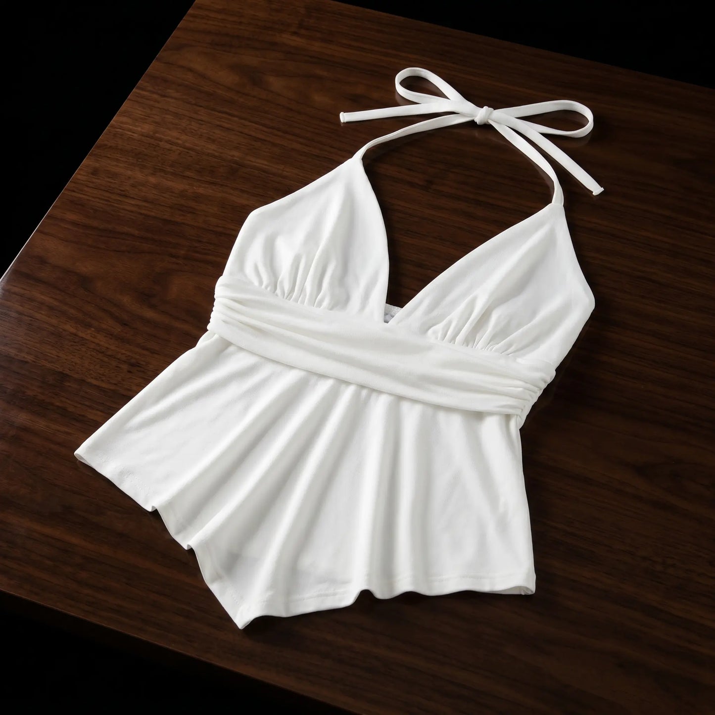 Off-white viscose-elastane blend halter neck camisole with a deep V-neck, cinched peplum waistband, and asymmetrical handkerchief hem, presented flat lay on a wooden table.