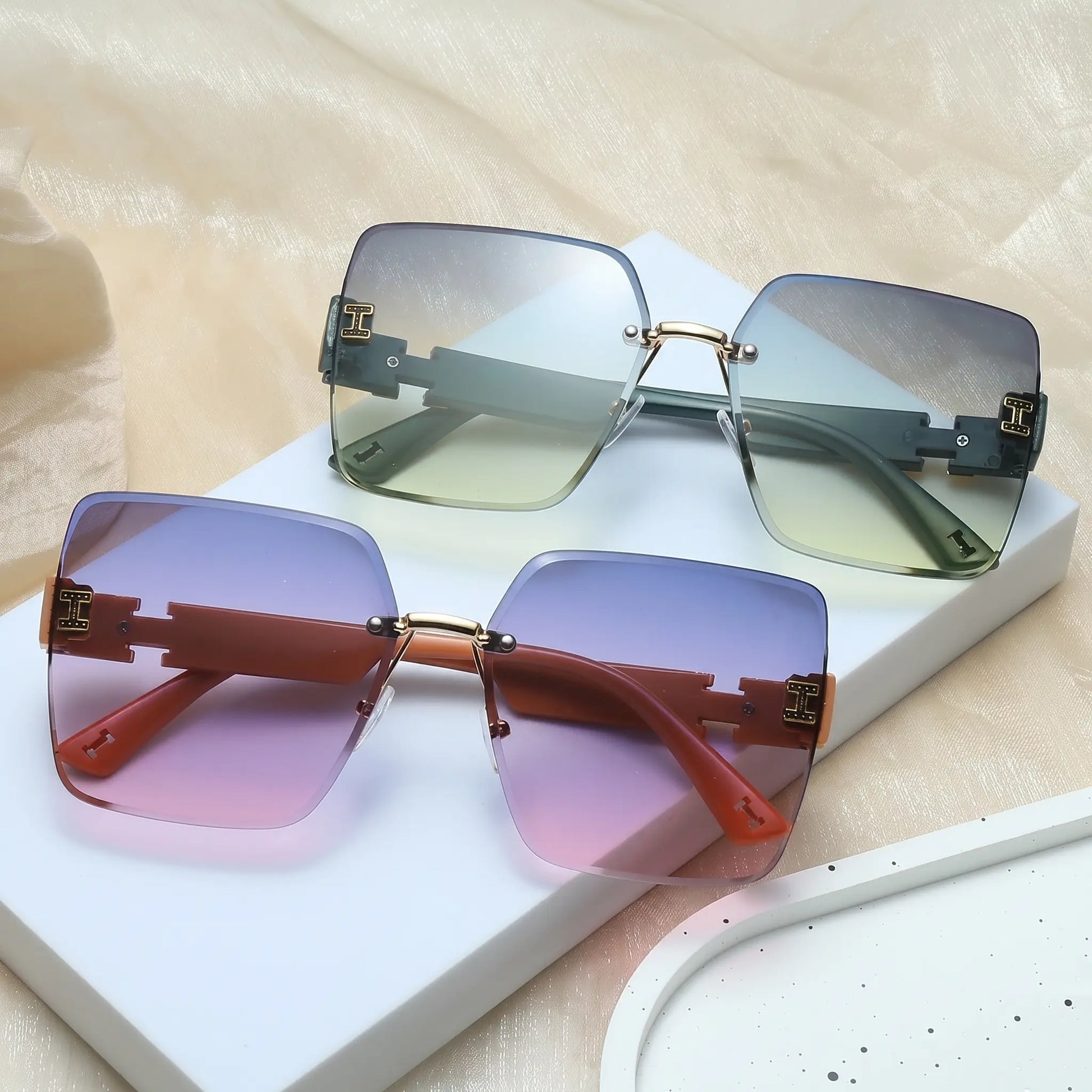 Two pairs of Aethel oversized square rimless sunglasses with gradient lenses and gold 'H' logo temple accents, displayed from a three-quarter angle.