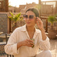 Woman wearing Aethel oversized square gradient sunglasses with gold 'H' logo accents, relaxing on a sun-drenched terrace with a drink.