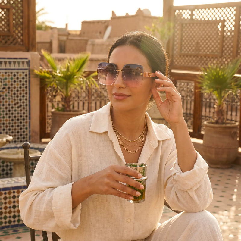 Woman wearing Aethel oversized square gradient sunglasses with gold 'H' logo accents, relaxing on a sun-drenched terrace with a drink.