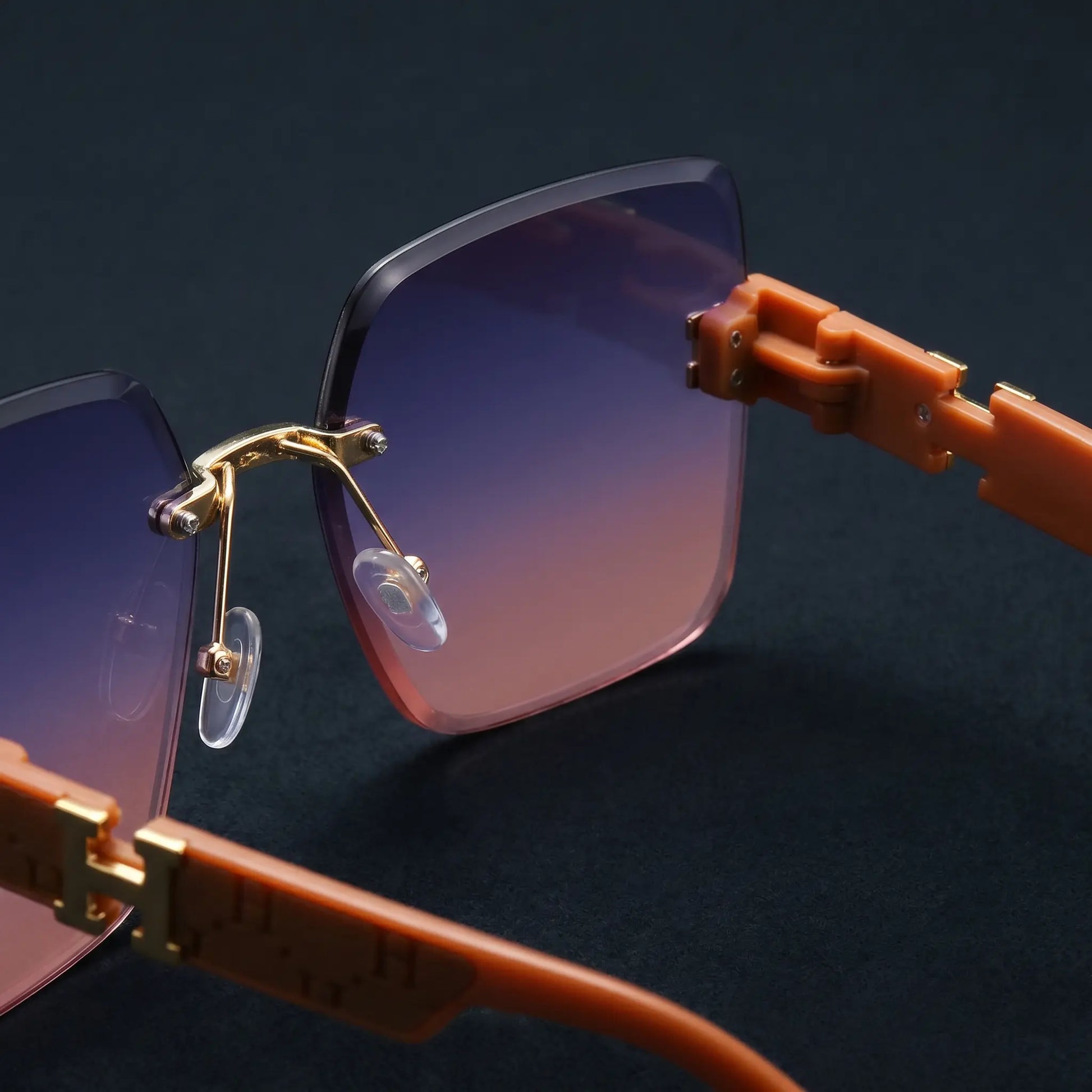 Oversized square sunglasses with gradient blue-pink lenses and distinctive gold-tone 'H' logo accents on the burnt orange temples, shown from a three-quarter angle.