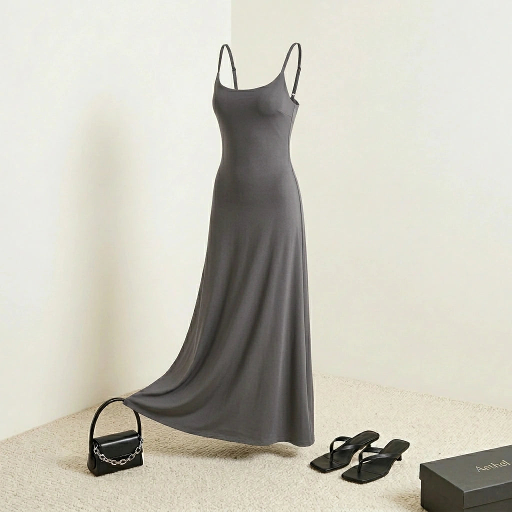 Aethel sleek maxi dress in charcoal grey with adjustable straps, styled with a black handbag and strappy sandals in a studio setting.