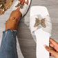 A pair of Aethel white flat slide sandals adorned with a gold-tone butterfly embellishment featuring crystal accents, styled on a model's feet with blue jeans and an anklet on a wooden floor.