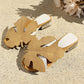 A pair of Aevana bohemian flat slide sandals in warm khaki, featuring oversized leaf embellishments and crystal-adorned low block heels, presented in a styled flat lay on a textured surface.