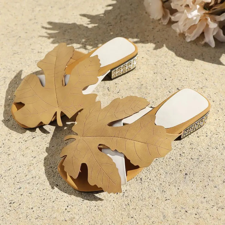 A pair of Aevana bohemian flat slide sandals in warm khaki, featuring oversized leaf embellishments and crystal-adorned low block heels, presented in a styled flat lay on a textured surface.