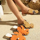 Aevana bohemian flat slides featuring oversized leaf embellishments in vibrant orange and warm khaki, shown worn by a model on a textured outdoor surface with crystal-embellished low block heels.