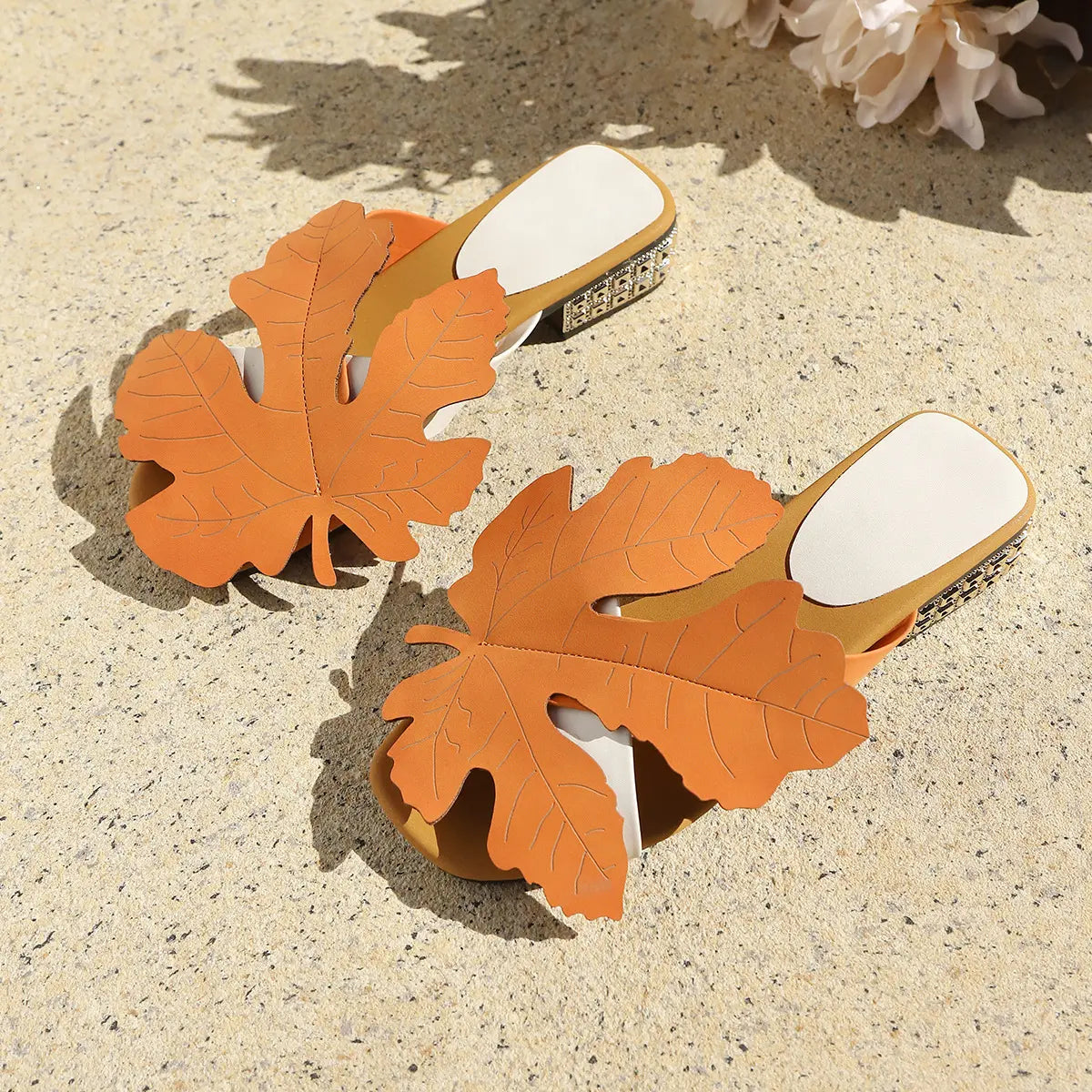 Aevana vibrant orange leaf-embellished flat sandals with crystal-accented low block heels, styled on a textured outdoor surface with natural shadows and flowers.
