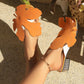 Model wearing vibrant orange leaf-embellished flat sandals with crystal-adorned low block heels, styled with an anklet in a sunny outdoor setting.