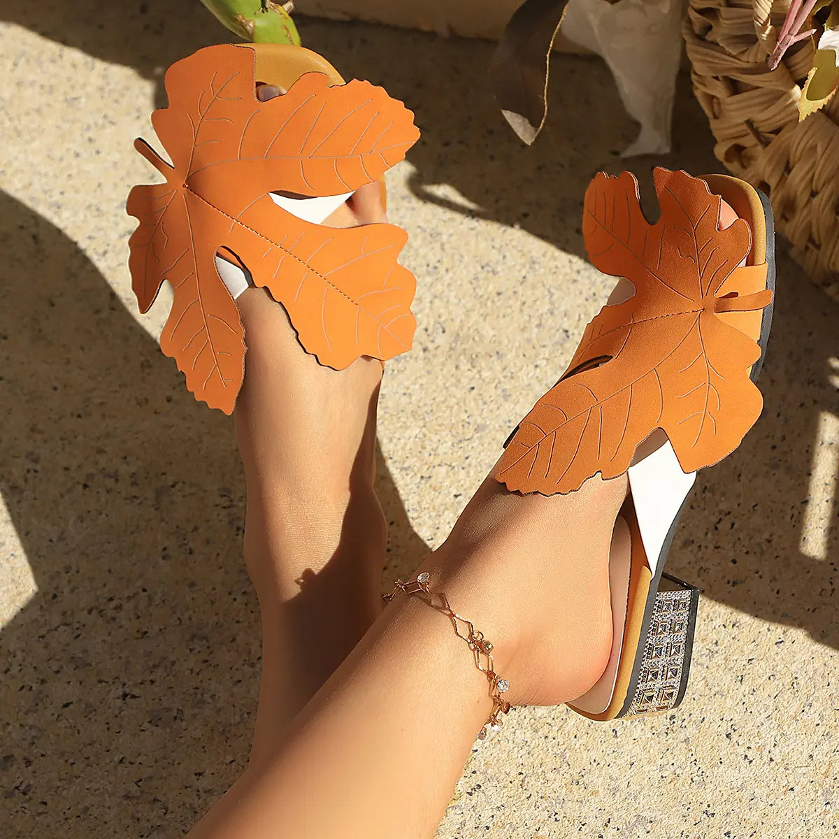 Model wearing vibrant orange leaf-embellished flat sandals with crystal-adorned low block heels, styled with an anklet in a sunny outdoor setting.