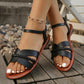Black Aevoria flat sandals with braided crossover straps and an elasticated ankle strap, worn by a model on a wooden deck with gravel.
