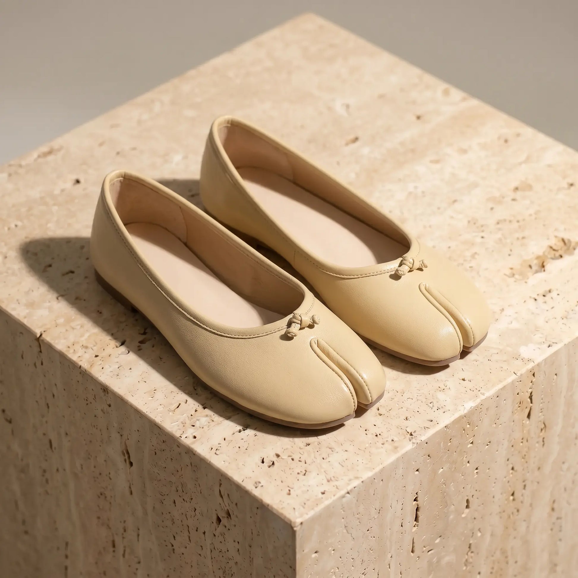Pair of soft apricot vegan leather split-toe ballet flats with delicate bows, displayed from a three-quarter view on a textured stone block.