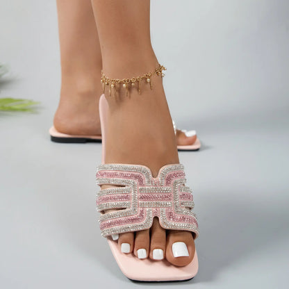 Close-up front view of Ayori pink rhinestone flat slide sandals, showcasing the embellished single band strap, square toe, and padded footbed on a foot with white toenails.