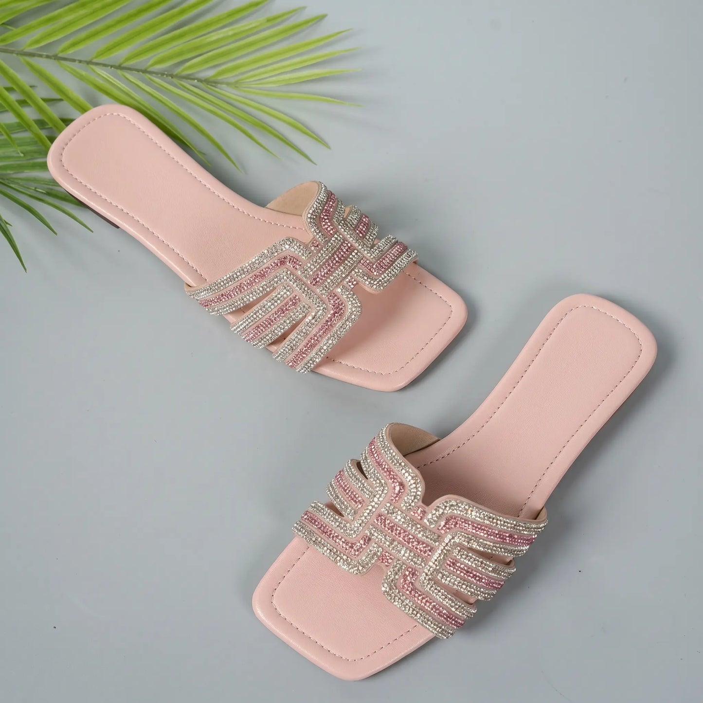 Ayori rhinestone flat slides in blush pink, featuring a chic square toe and a single embellished band strap, displayed from a three-quarter angle with a decorative palm leaf accent.