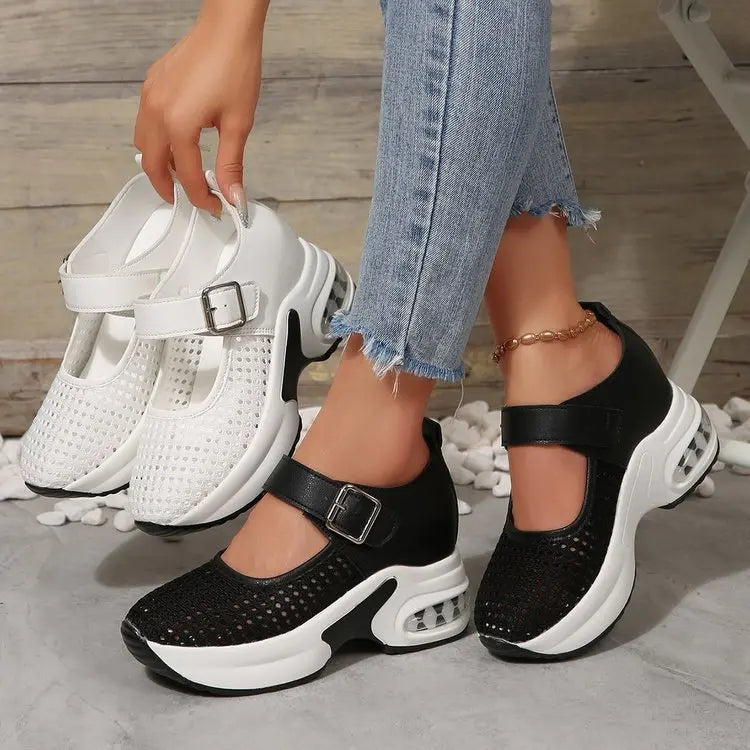 Women's breathable platform sneakers in black and white, featuring a perforated mesh upper, adjustable buckle strap, and visible air cushion heel. Shown on a model's feet with distressed denim jeans.