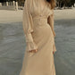 Woman wearing a warm beige chiffon midi dress with a romantic V-neckline, fitted waist, and voluminous bishop sleeves with pearl button cuffs, standing by the water on a beach.