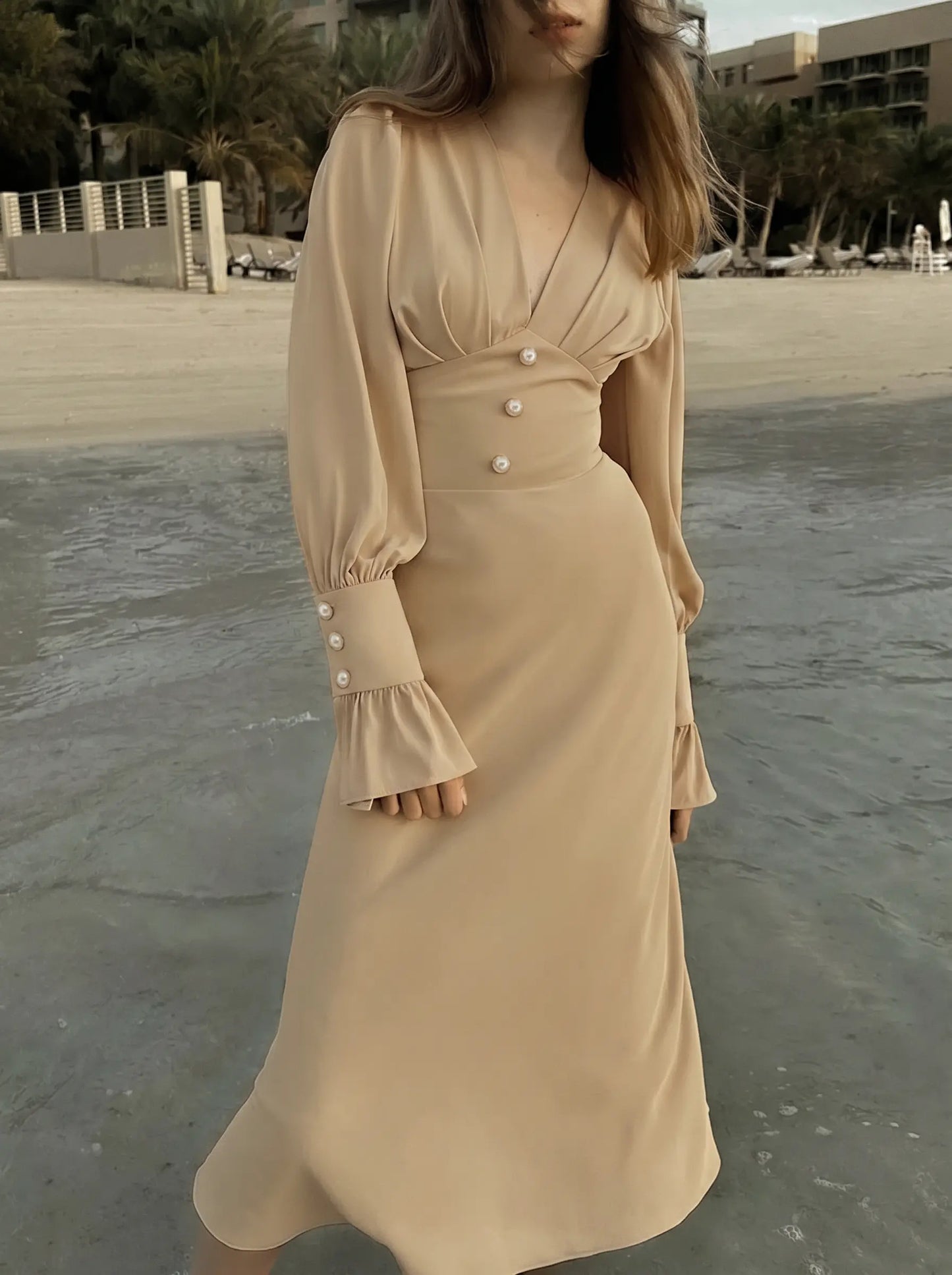 Woman wearing a warm beige chiffon midi dress with a romantic V-neckline, fitted waist, and voluminous bishop sleeves with pearl button cuffs, standing by the water on a beach.