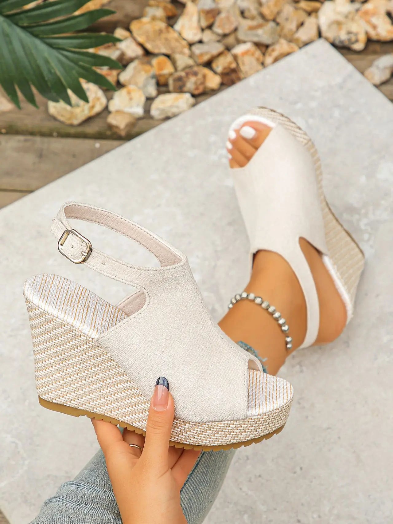 Beige espadrille wedge sandals worn on a foot, showcasing the peep-toe design, jute-wrapped wedge, and adjustable ankle strap, with another sandal held in hand. Features a textured suede-like upper and natural bohemian styling.