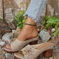 Beige peep-toe wedge sandals with layered floral applique and perforated straps, worn with denim jeans in an outdoor, natural setting.