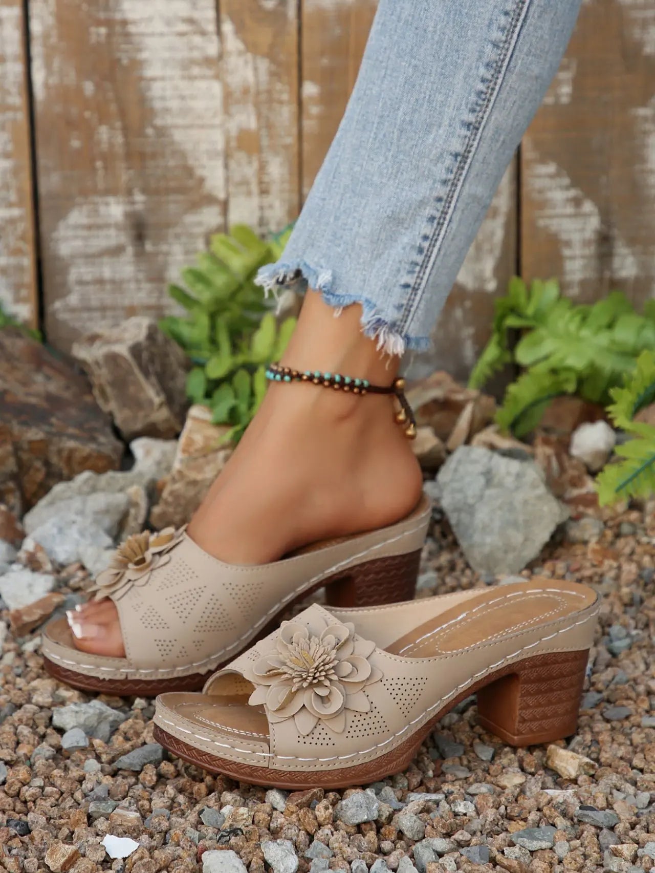 Beige peep-toe wedge sandals with layered floral applique and perforated straps, worn with denim jeans in an outdoor, natural setting.