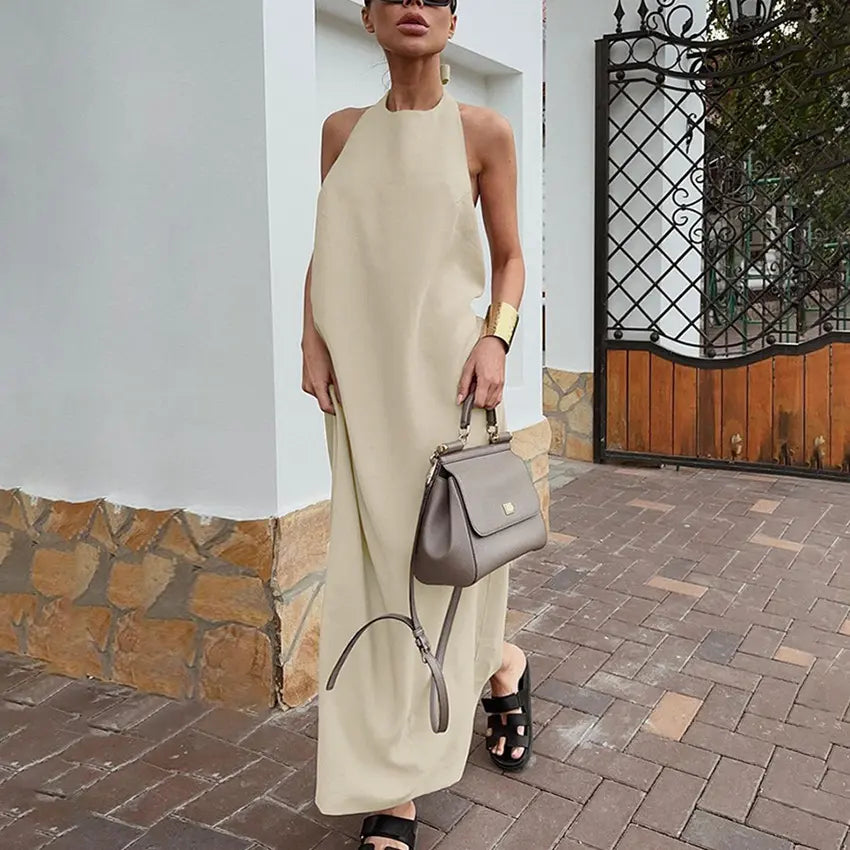 A woman models a flowing, sleeveless halter neck maxi dress in a light beige cotton-linen blend, styled with a handbag and sandals in an outdoor setting.