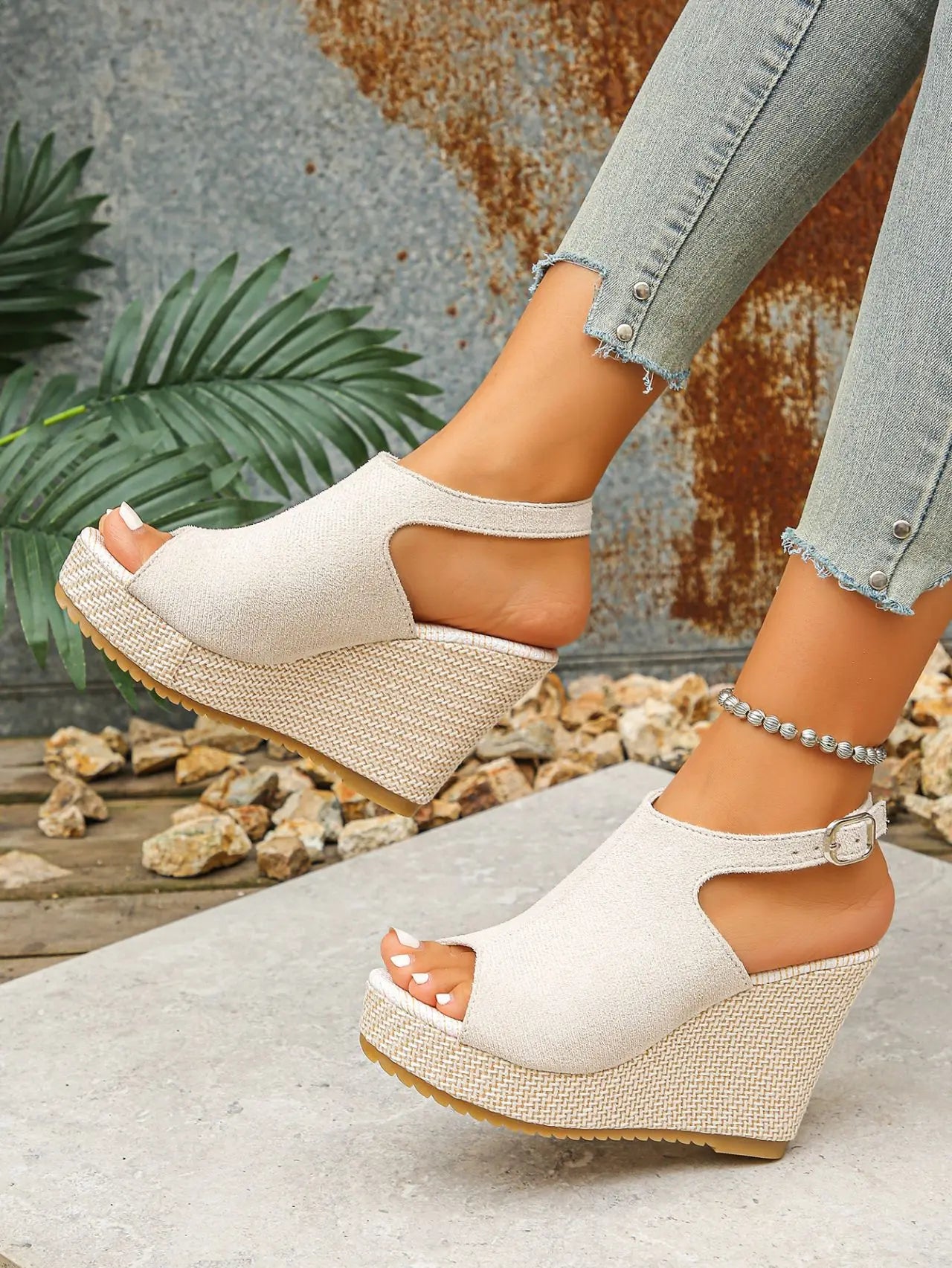 A woman's feet wearing beige peep-toe espadrille wedge sandals with jute-wrapped heels, paired with distressed denim jeans and an anklet, in a natural outdoor setting.