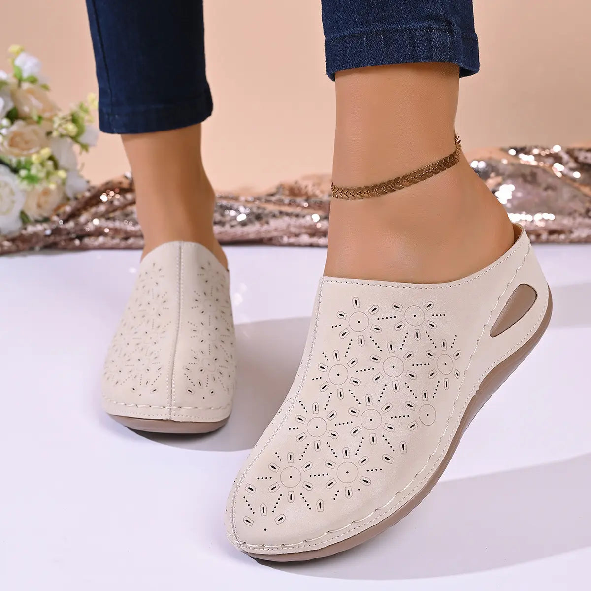 Close-up of beige perforated wedge mules on a model's feet, showcasing intricate floral laser-cut patterns, white stitching, and a low wedge heel.