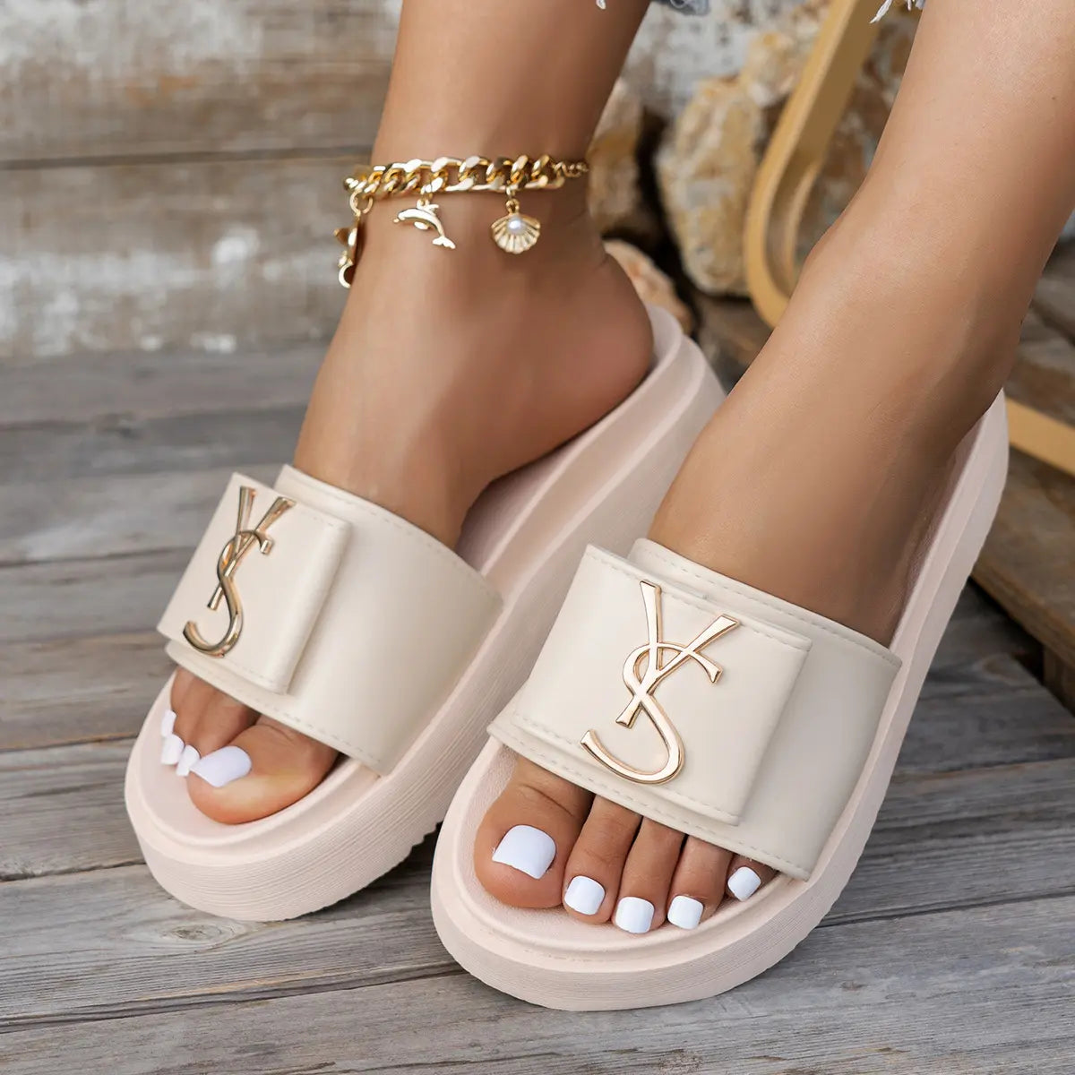 Women's light beige platform slides featuring a wide strap with a polished gold-tone metallic logo accent and a comfortable, sculpted sole, worn on feet with white nail polish.