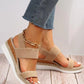 Close-up of beige woven espadrille wedge sandals with a gold chain accent on the ankle, worn with blue jeans on a wooden floor.
