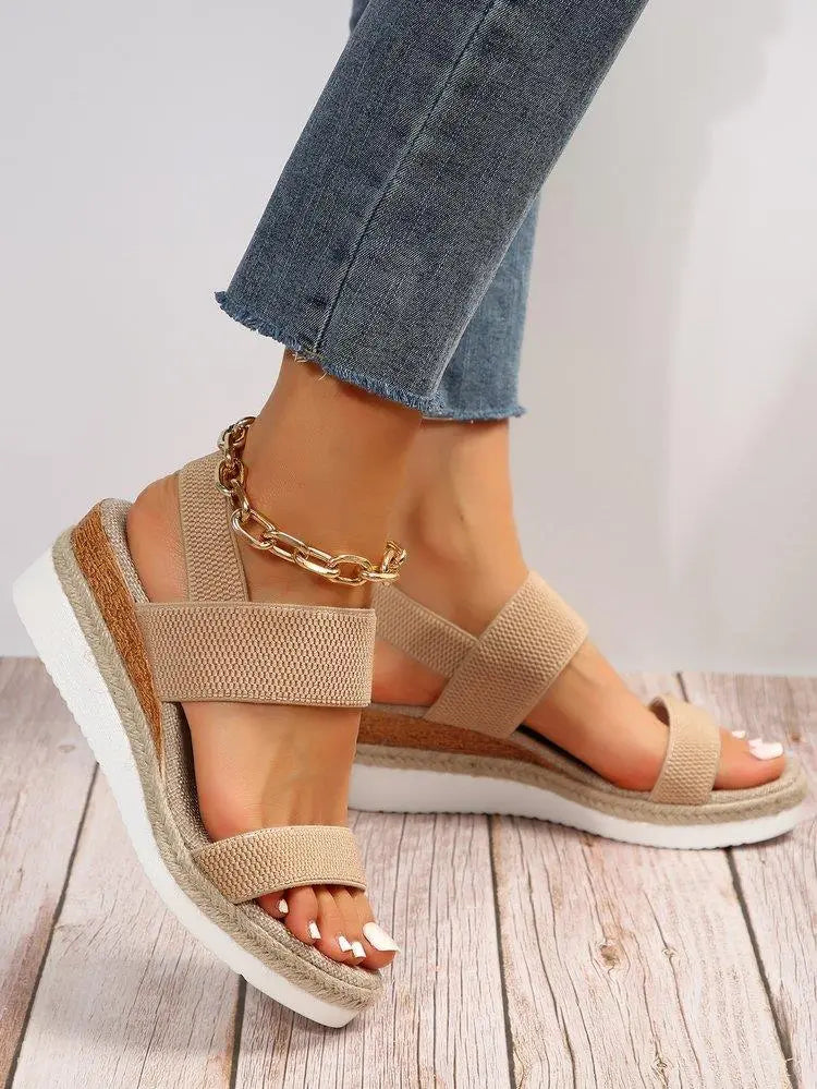 Close-up of beige woven espadrille wedge sandals with a gold chain accent on the ankle, worn with blue jeans on a wooden floor.