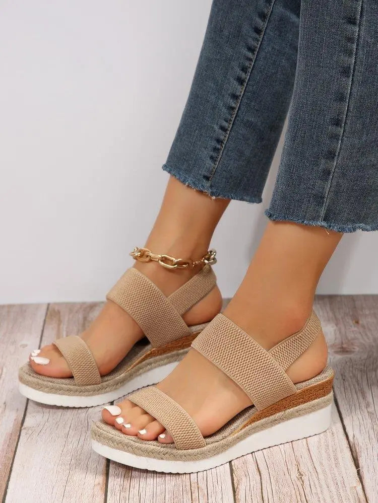 Women's beige woven espadrille wedge sandals with a fish-mouth open toe and elasticated slingback strap, styled with denim jeans on a wooden floor.