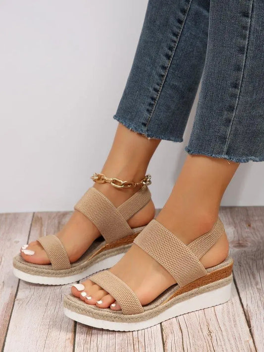 Women's beige woven espadrille wedge sandals with a fish-mouth open toe and elasticated slingback strap, styled with denim jeans on a wooden floor.
