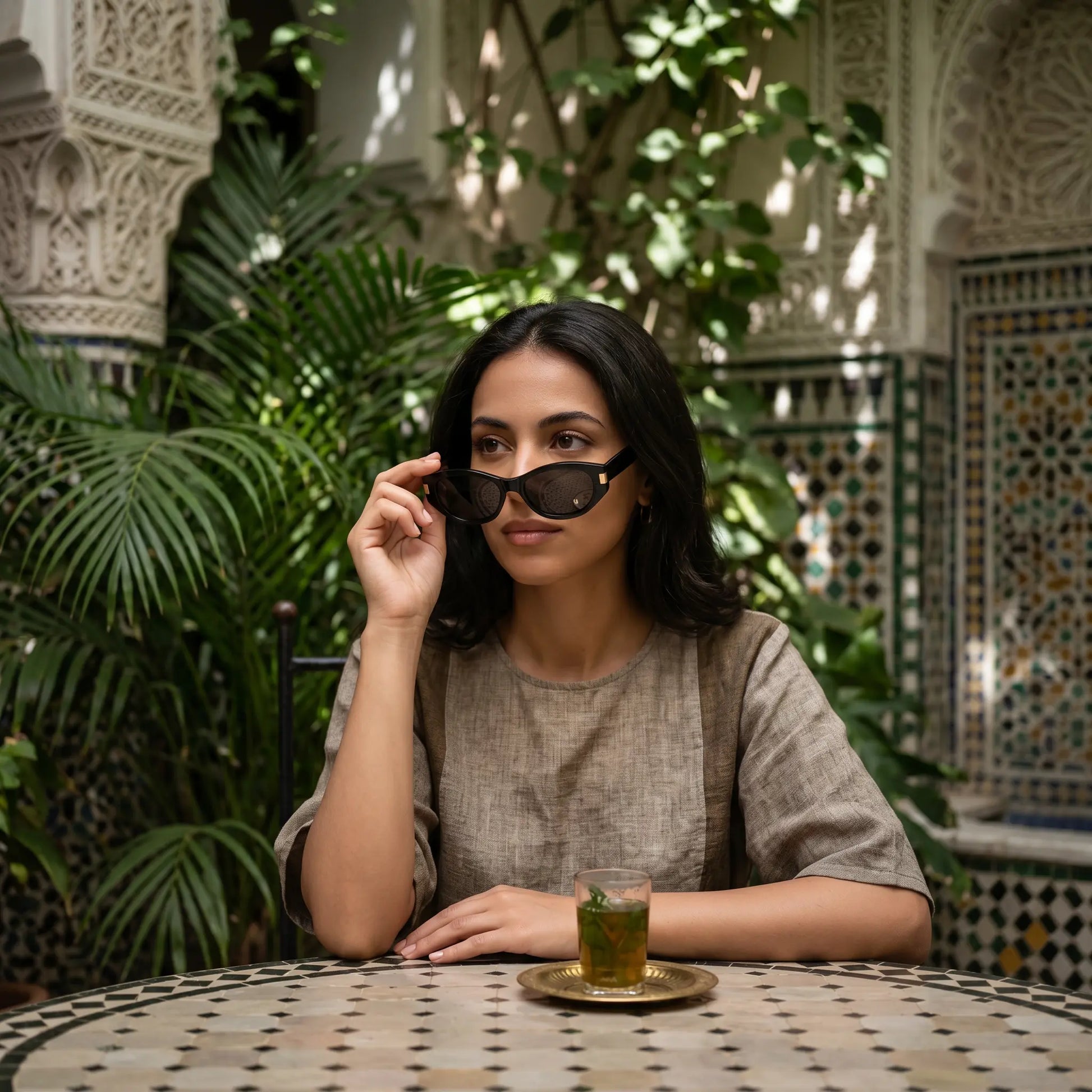 Elegant woman adjusting black acetate cat-eye sunglasses with gold-tone accents, seated at a mosaic table in a lush, traditional Riad setting.