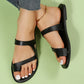 Elegant black asymmetrical strap flat slide sandals for women, featuring an open-toe design and white contrast stitching detail on the sole, shown worn on feet with denim jeans.