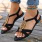 Black bohemian flat sandals with a pointed toe, featuring interwoven straps adorned with gold beads, carved wood accents, and a central gold flower charm. Worn by a model with distressed denim jeans on a weathered wooden deck.