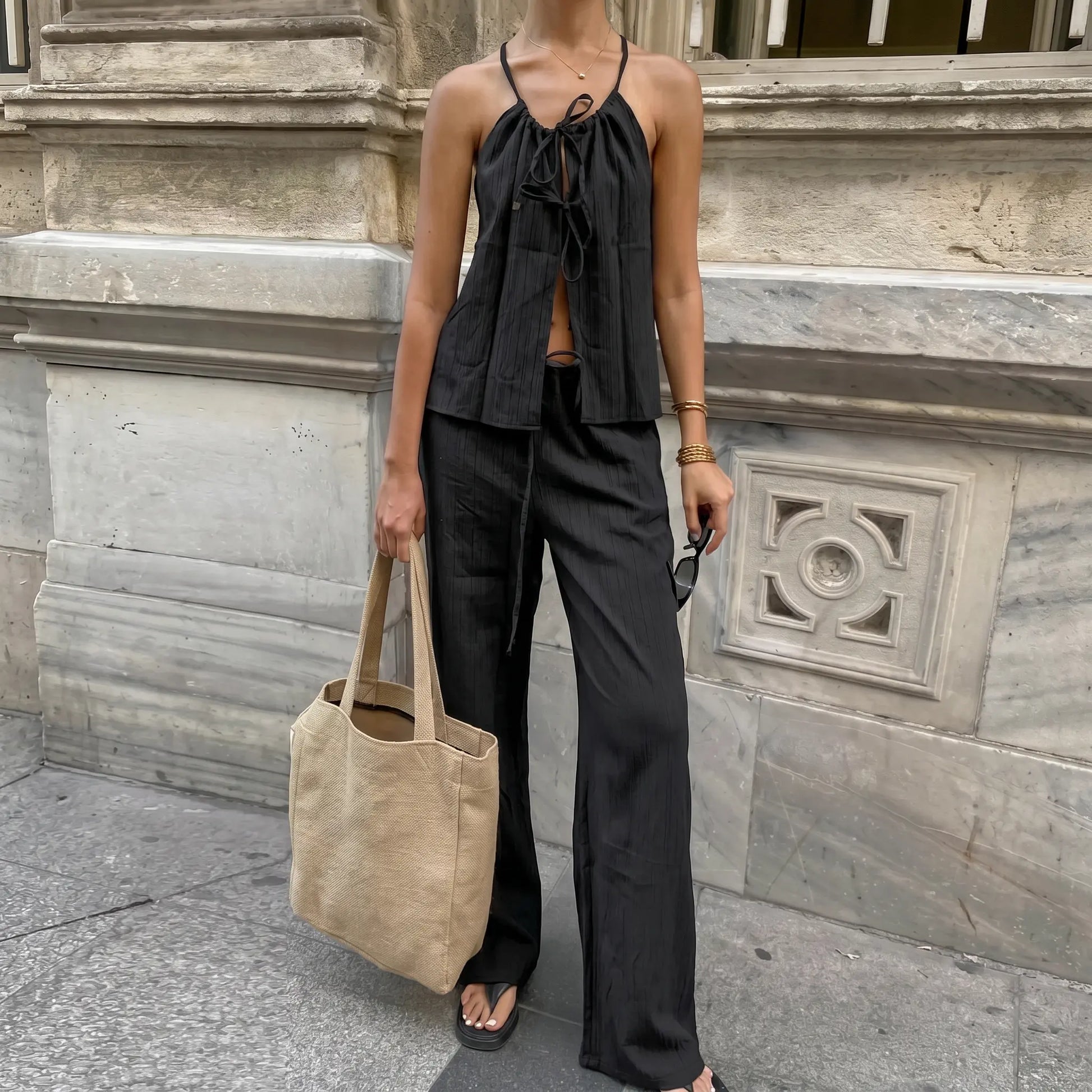 A woman models a black bohemian linen halter neck top with adjustable tie and matching wide-leg trousers in an urban outdoor setting, showcasing an elegant summer outfit.