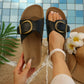 Black slip-on slide sandals with antique brass buckles shown on a foot and held by a hand, set against a poolside lifestyle background with a straw hat and tropical flower. Features a cork footbed and open-toe design.
